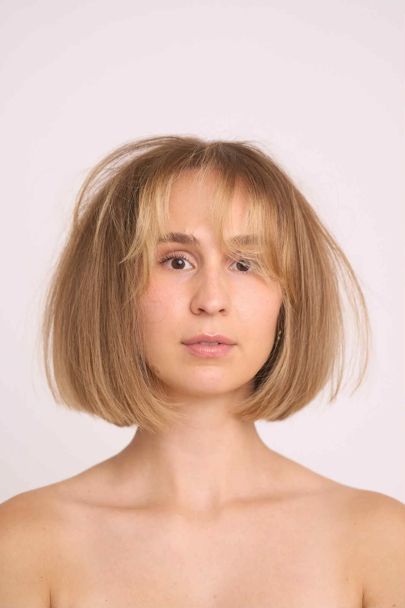 Close-up of a young woman with shoulder-length, tousled light brown hair, fair skin, and light makeup, against a plain white background.