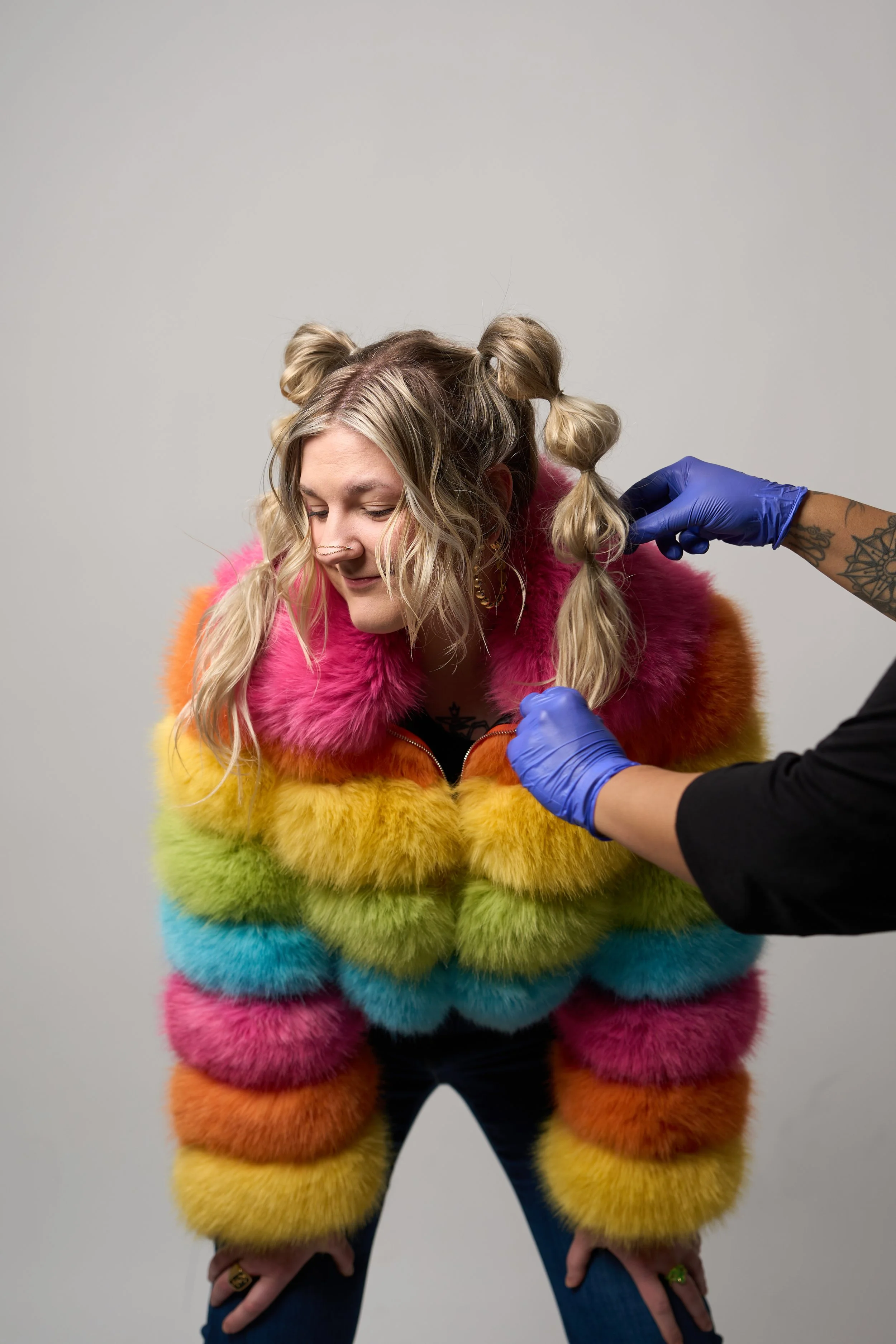 A woman with blonde curly hair styled in playful buns is getting her hair done by a person wearing blue gloves. She is wearing a colorful, furry, rainbow-striped jacket and is smiling.