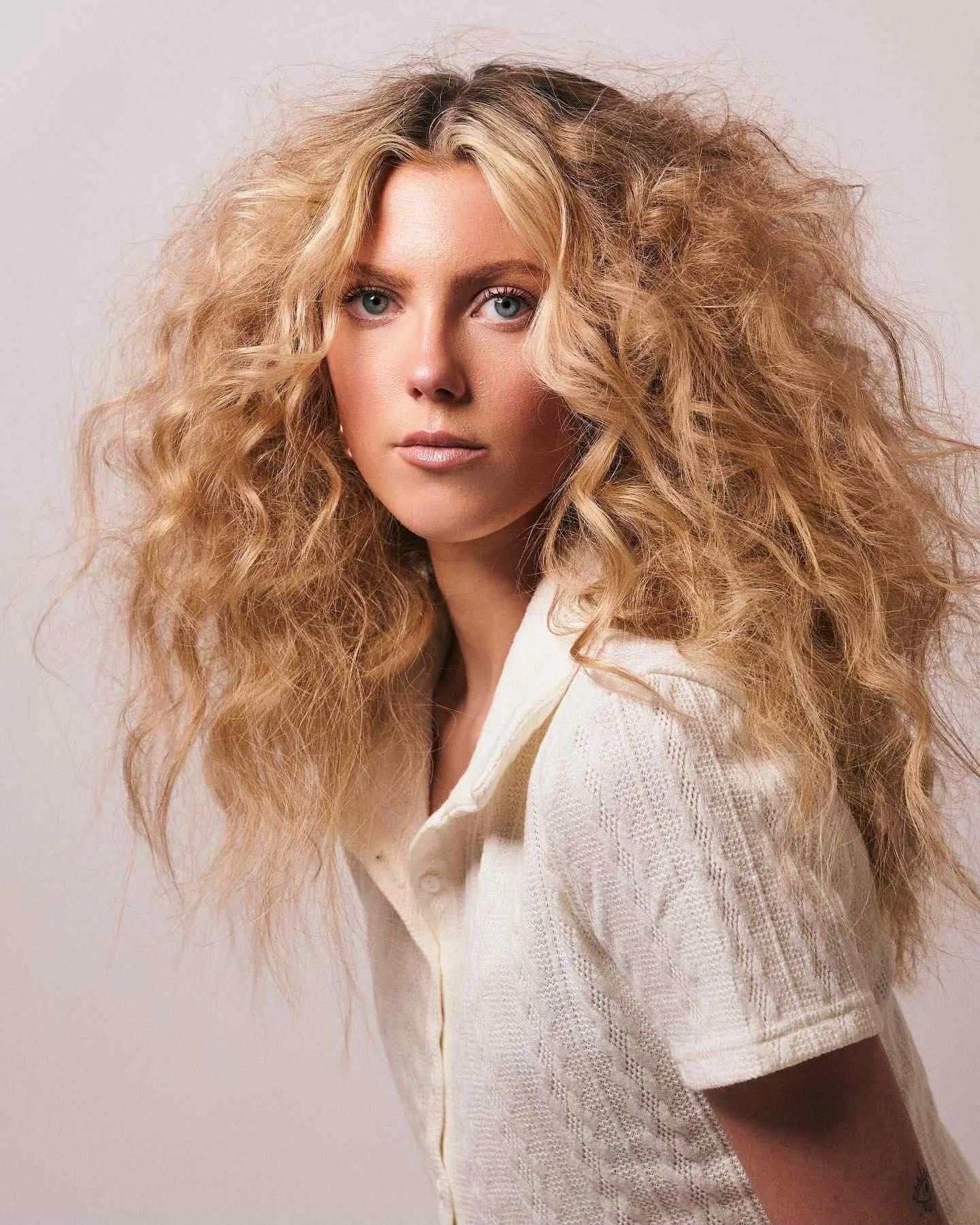A woman with long, curly blonde hair wearing a white shirt against a plain background.