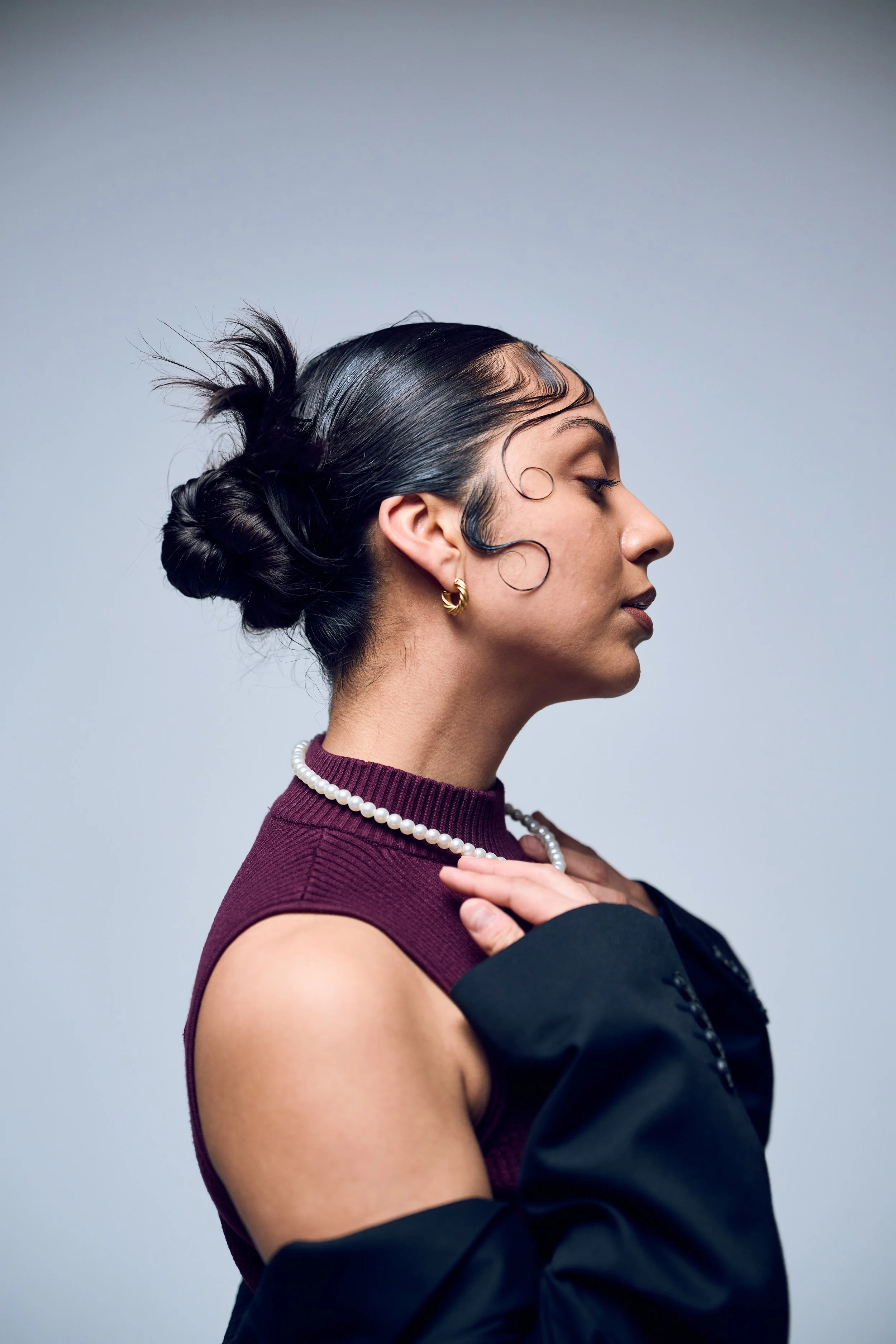 A woman with dark hair styled in a bun and intricate curling on the side, wearing pearl necklace, gold hoop earrings, a sleeveless maroon top, and a black jacket draped over her shoulders, posing in profile against a plain gray background.
