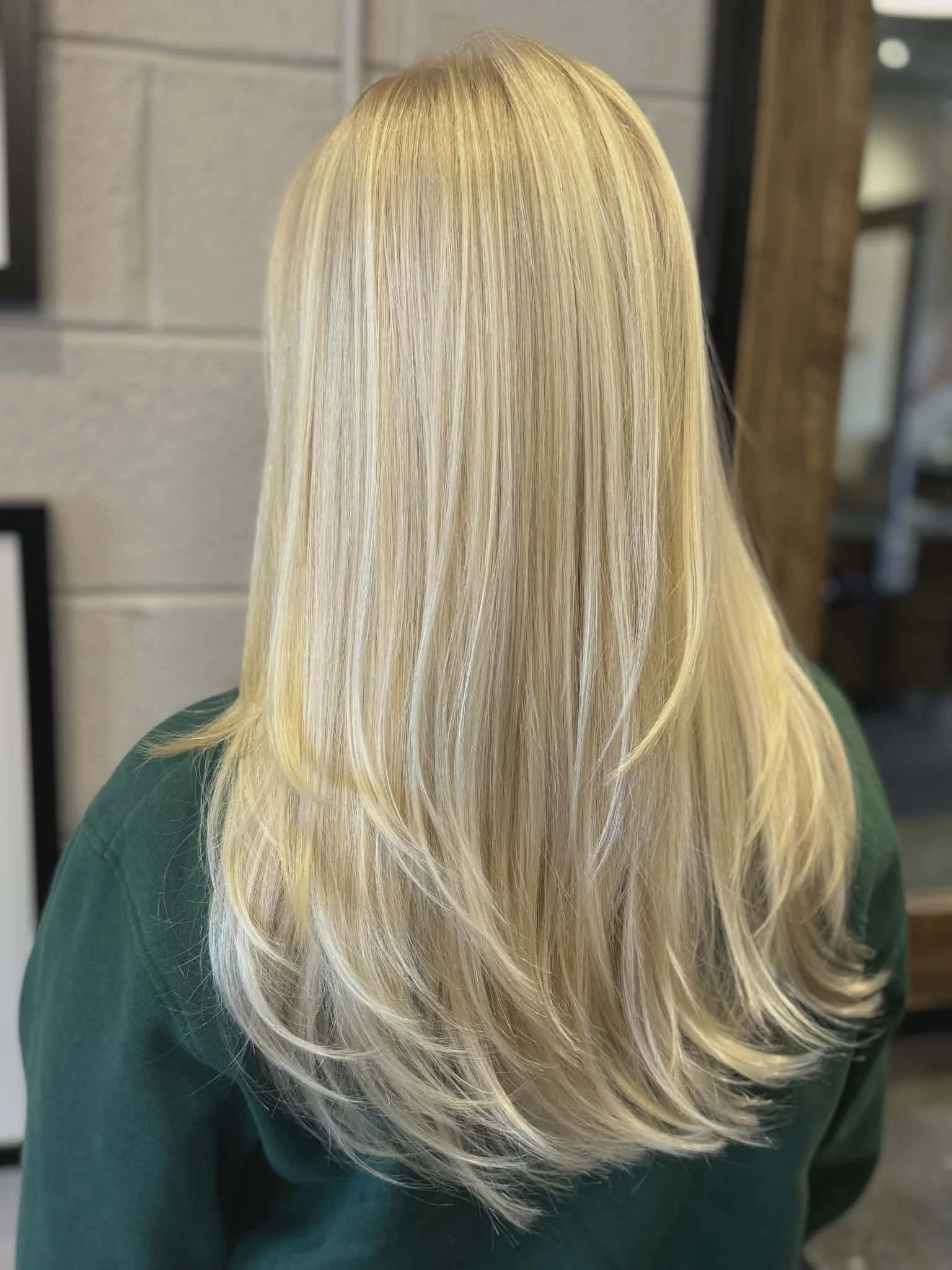 Back view of a woman with long, straight blonde hair wearing a green top, in an indoor setting.