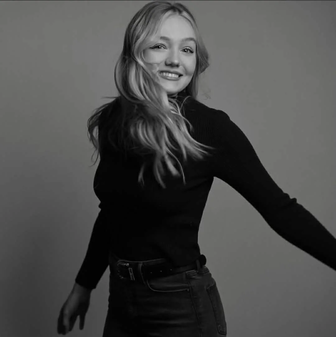 Black and white portrait of a young woman with long, wavy hair, smiling and looking at the camera, wearing a long-sleeve top and jeans, against a plain gray background.