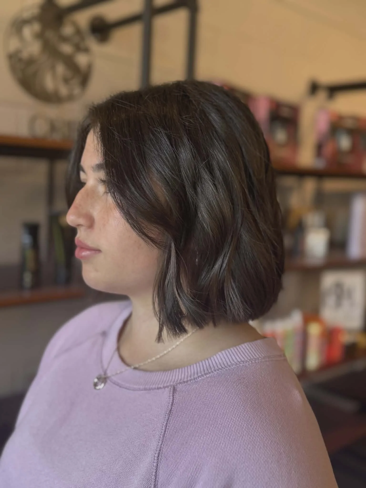 Side profile of a woman with short, wavy, dark brown hair wearing a light purple shirt and a necklace, in a cozy indoor setting with shelves and decorative items in the background.