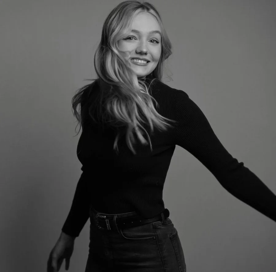 Black and white photo of a young woman with long wavy hair, smiling and looking at the camera, wearing a long-sleeve top and jeans, standing against a plain background.
