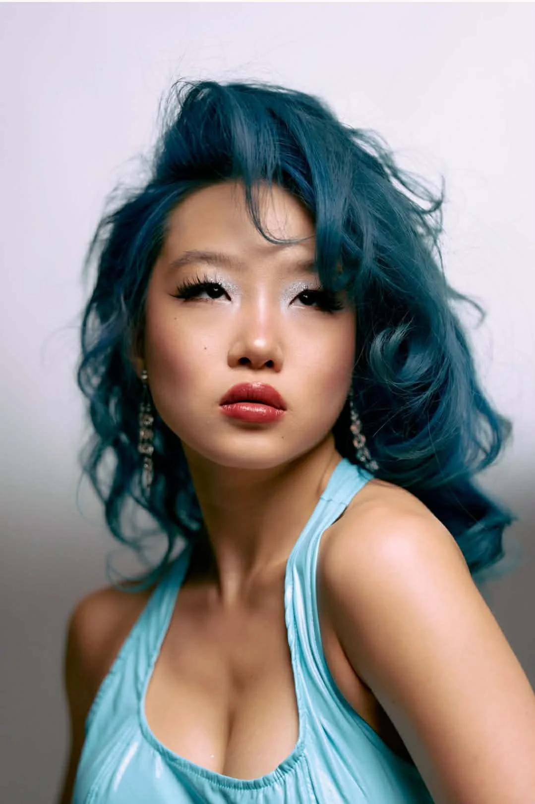 A young woman with vibrant blue, wavy hair takes a close-up portrait. She has dramatic makeup with silver eyeshadow, long eyelashes, and glossy lips. She is wearing a light blue sleeveless top and dangly earrings, against a neutral background.