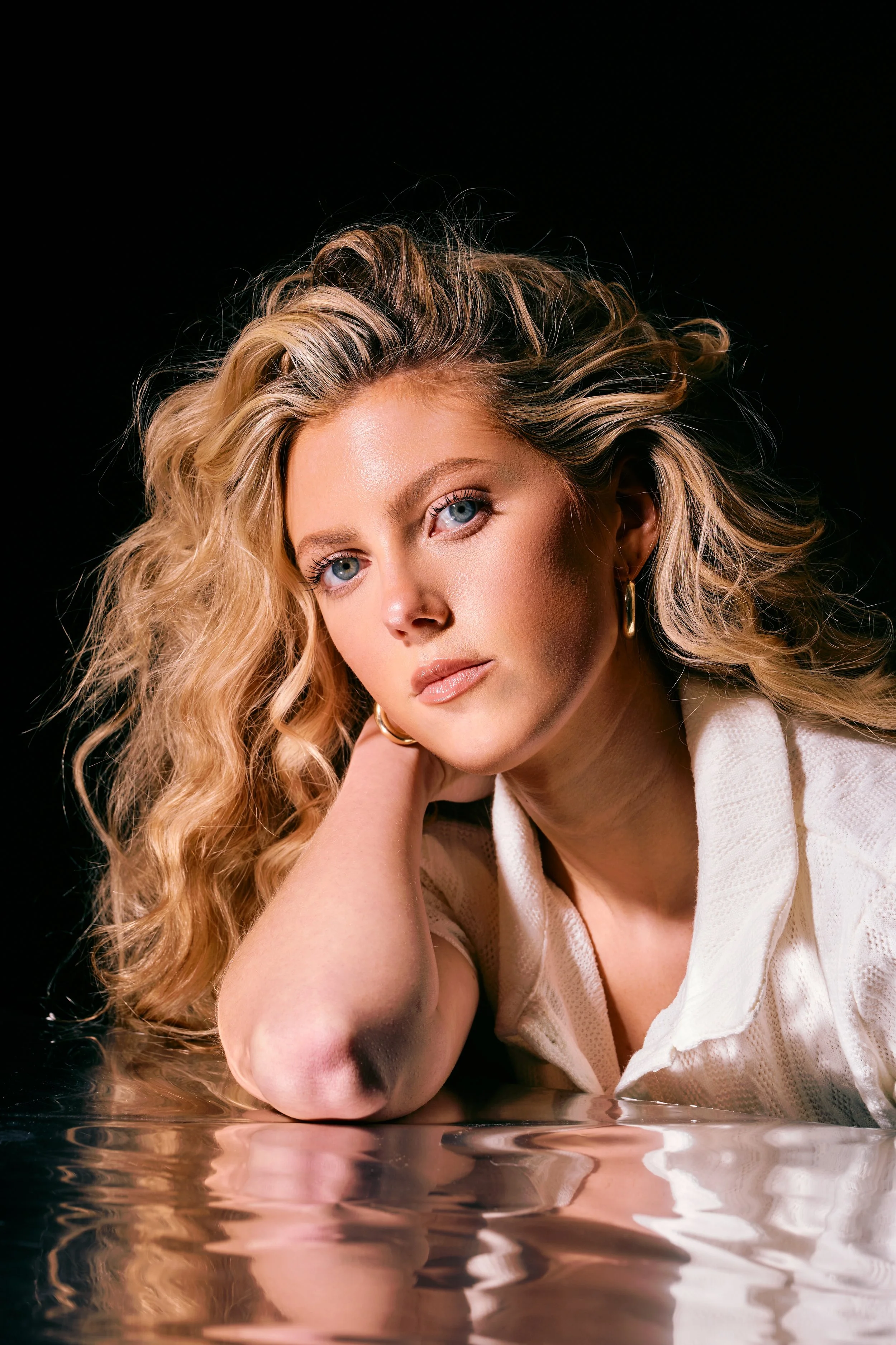 A woman with curly blonde hair and blue eyes rests her chin on her hand, leaning on a reflective surface, against a dark background.