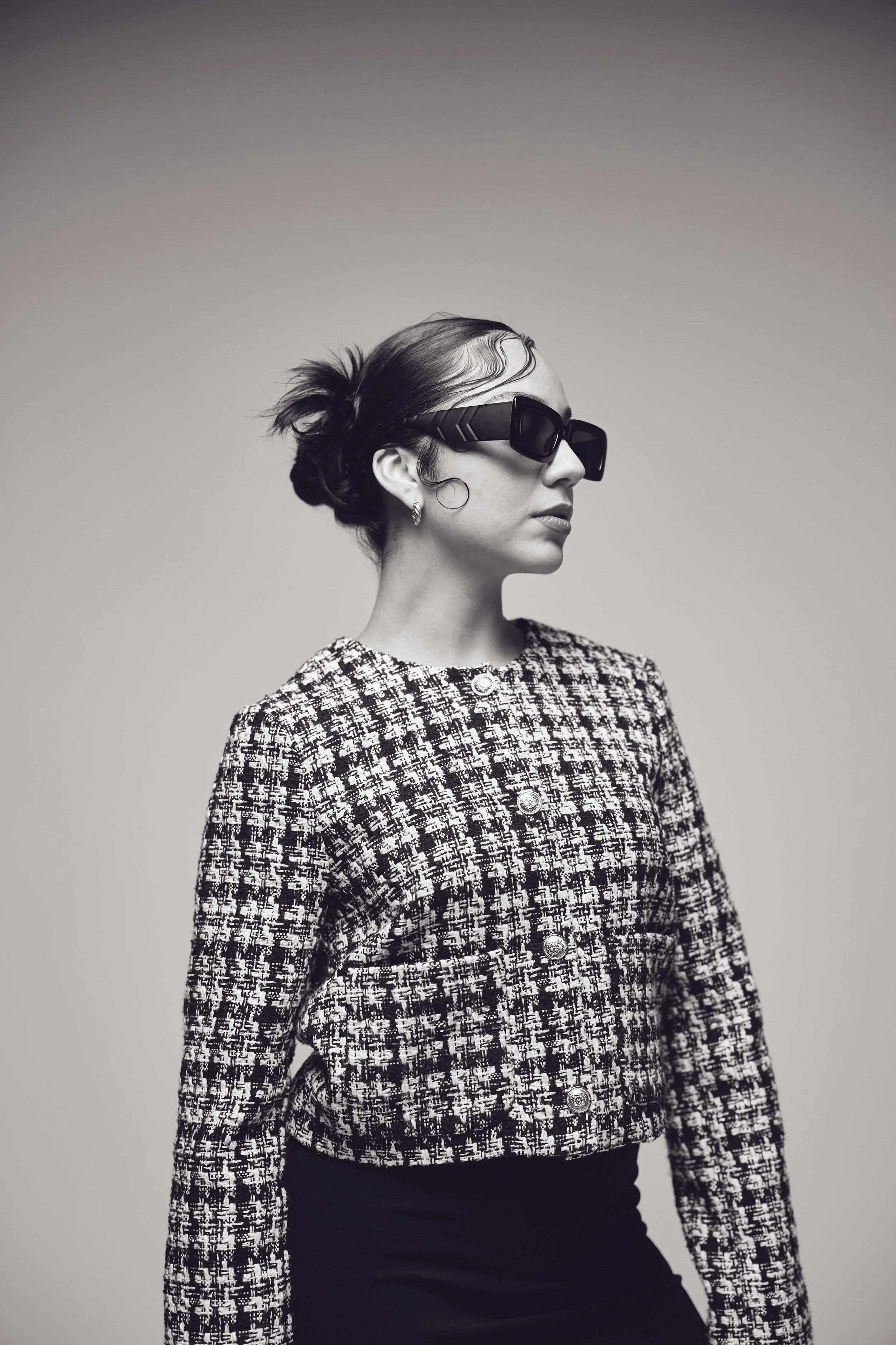 Black and white photo of a woman wearing large sunglasses, a patterned jacket, and a black skirt.