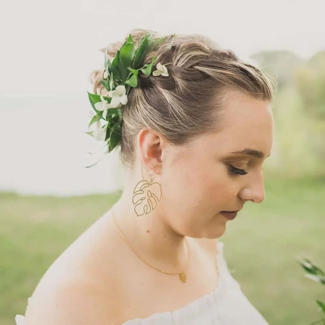 A woman with a floral crown and large leaf-shaped earring, her eyes closed, outdoors in a serene setting.
