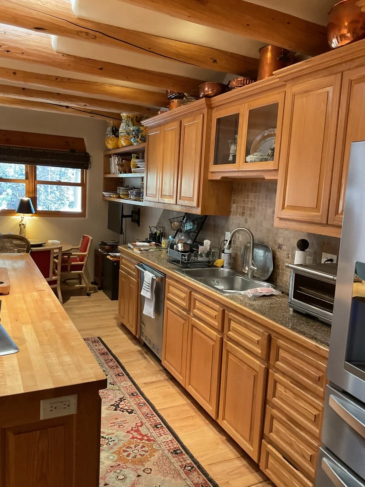 A cozy kitchen with wooden cabinets, a stone backsplash, and a wooden dining table. There is a window with a view of trees, and a corner nook with chairs and a lamp. The kitchen has various utensils, appliances, and decorative pottery on the shelves and top of the cabinets.