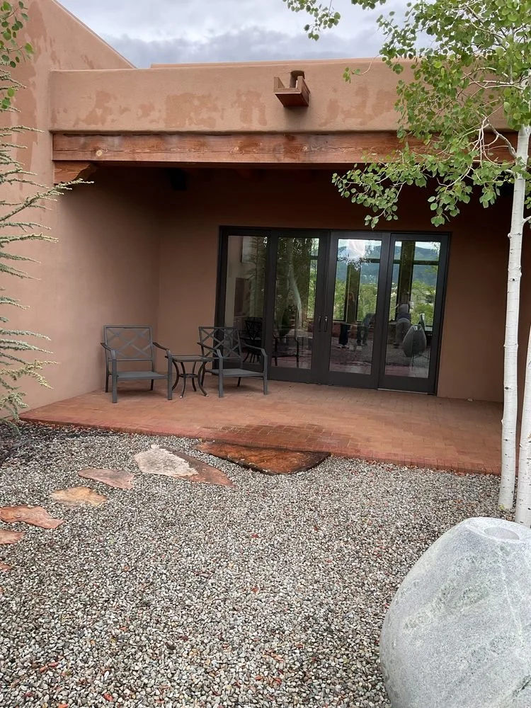 Exterior patio with two black chairs and a small table, sliding glass doors, gravel ground, and a tree on the right.