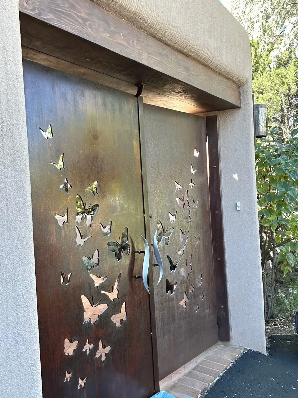 Metal gate with butterfly cutout designs, flanked by concrete wall and brick pathway. Green foliage to the right side.