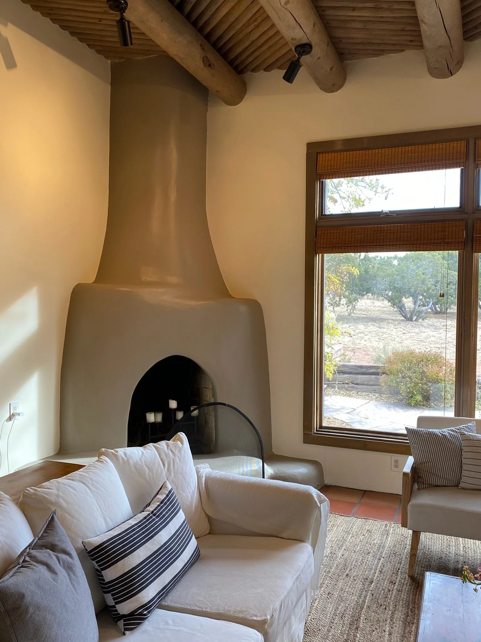 Living room with a cream-colored sofa, decorative pillows, a wood coffee table, large triangular fireplace with a chimney, and big window with wooden blinds showing an outdoor landscape with trees.