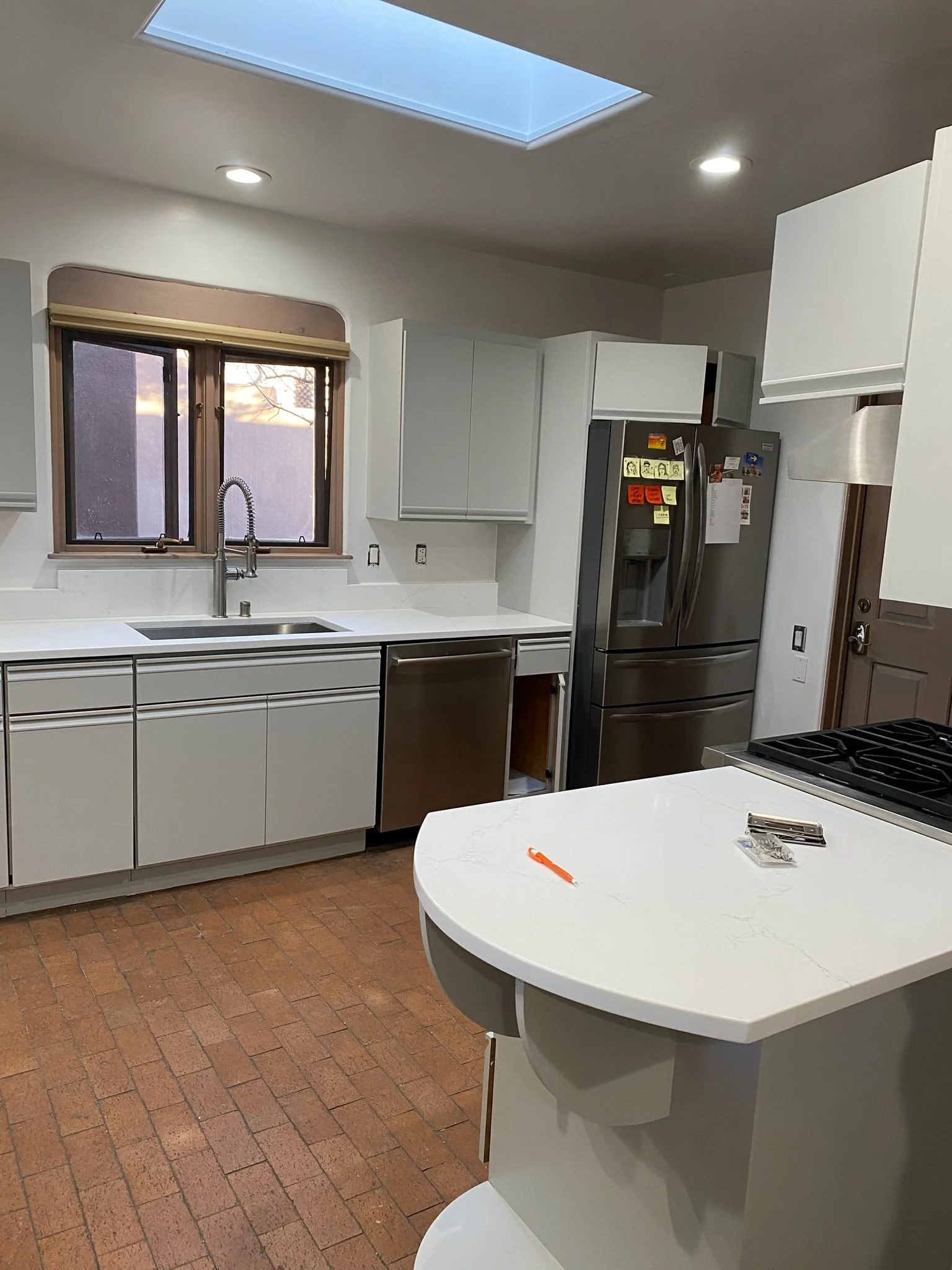 Kitchen under renovation with white cabinets, stainless steel appliances, a brick floor, an island with a white marble countertop, a window above the sink, and a skylight on the ceiling.