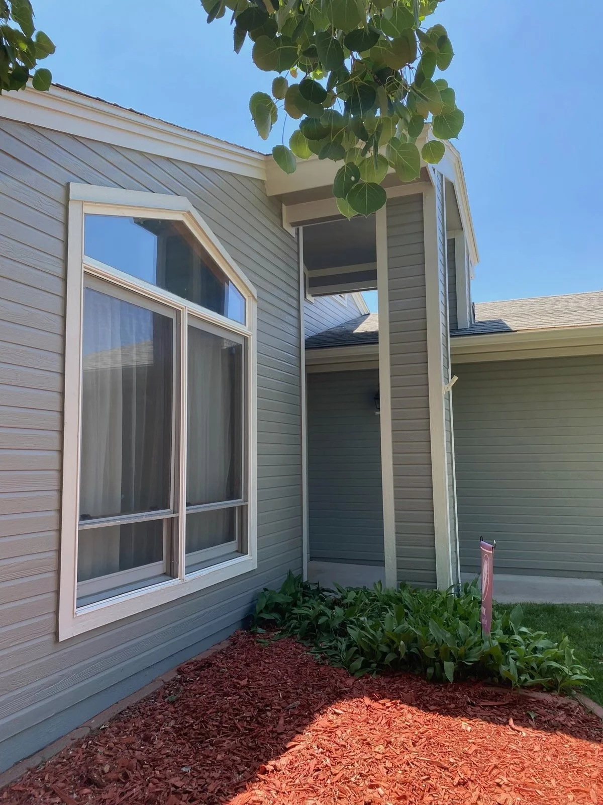Side view of a house with beige siding, a large window, and a small protruding covered porch area. There is a garden bed with green plants and reddish mulch in the foreground. A tree with large leaves is visible at the top.