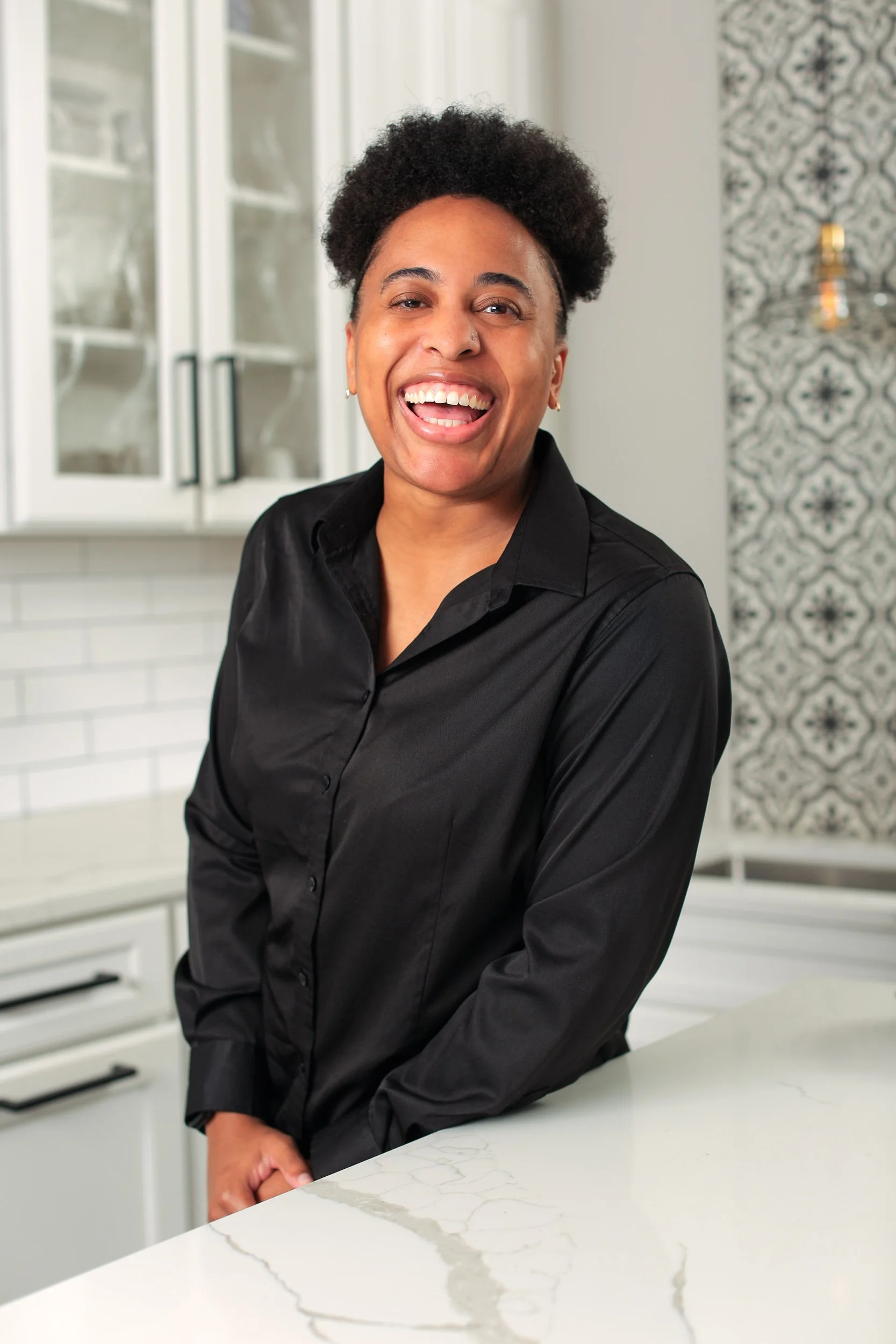 Smiling woman with short curly hair wearing a black blouse standing in a modern kitchen with white cabinets and decorative wallpaper.