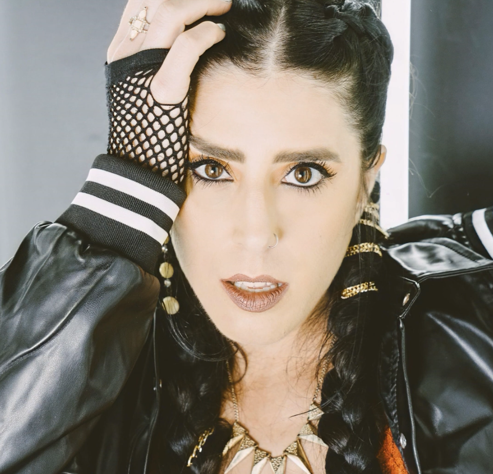 Close-up of a woman with dark braided hair, wearing a black leather jacket, fishnet glove on one hand, and gold accessories, looking directly into the camera with a surprised expression.