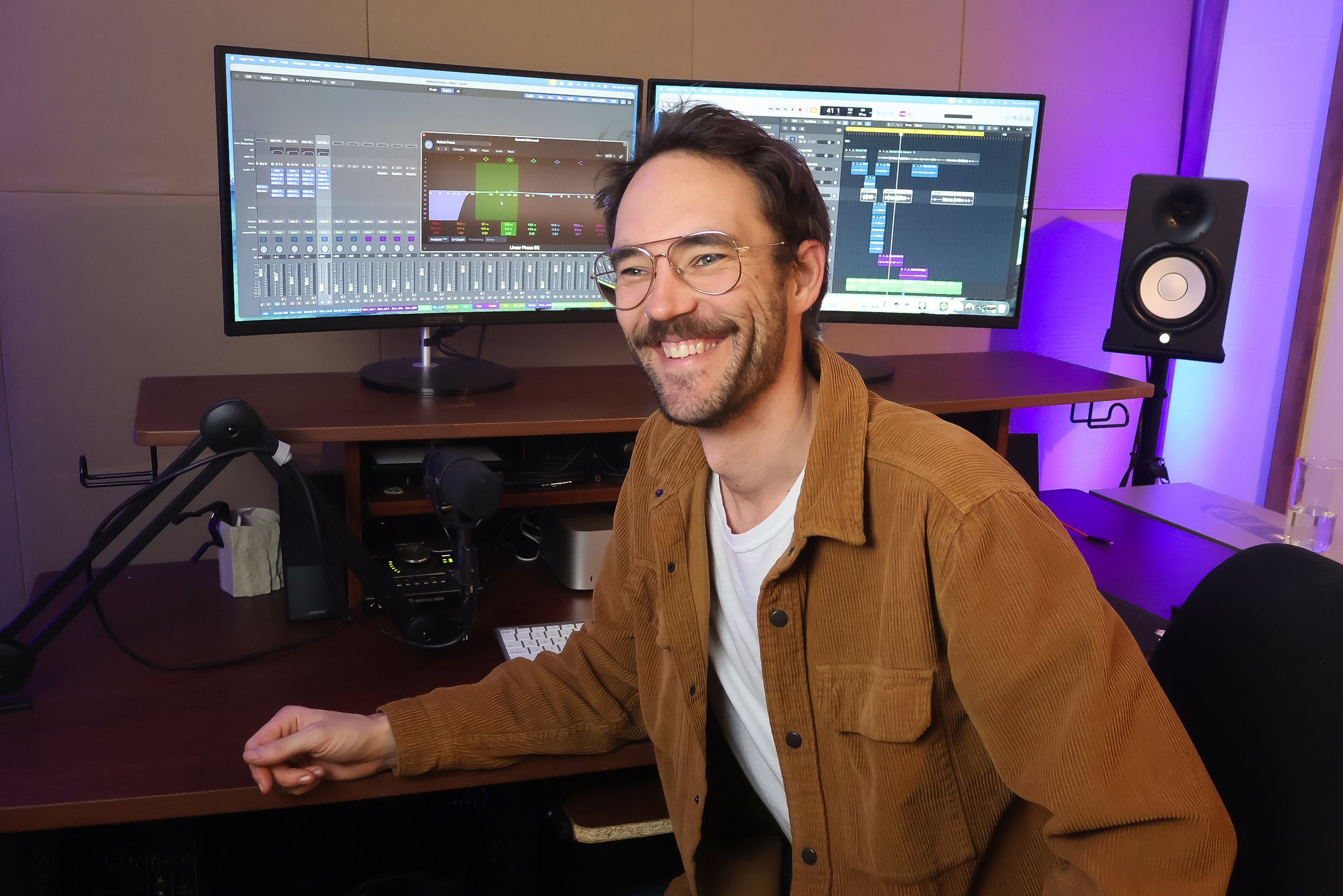 A man smiling sitting at a desk with two large computer monitors displaying music production software, a studio speaker, and various audio equipment.