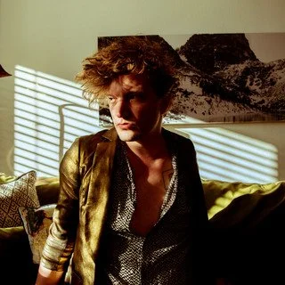 Young man with curly hair looking to the side in a room with sunlight and mountains artwork on the wall.