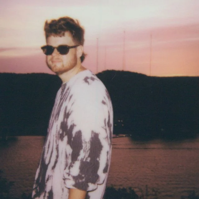 A young man with curly hair and sunglasses standing outdoors during sunset, wearing a tie-dye shirt with a river and hills in the background.