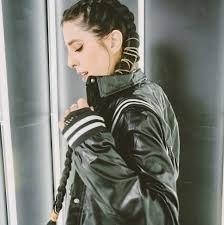 Young woman with braided hair wearing a black leather jacket, looking down and holding her hand to her collar, standing near a window.