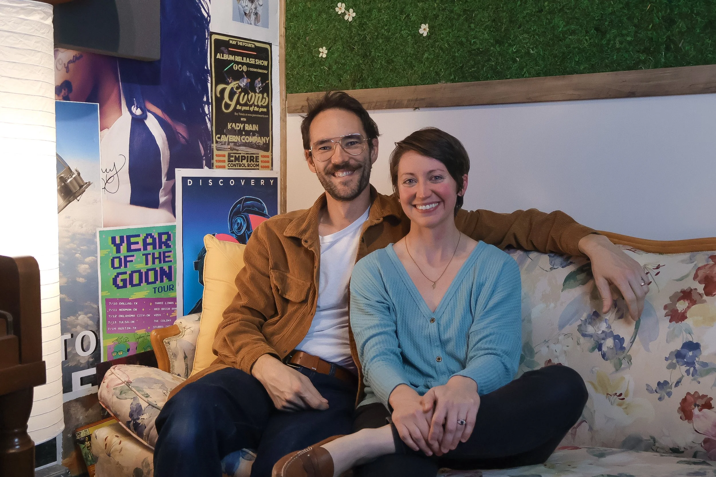 A smiling man and woman sitting on a floral-patterned couch, with the man's arm around the woman's shoulder. The background features posters, including one with a pixelated design and promotional text for a tour called "Year of the Goon."