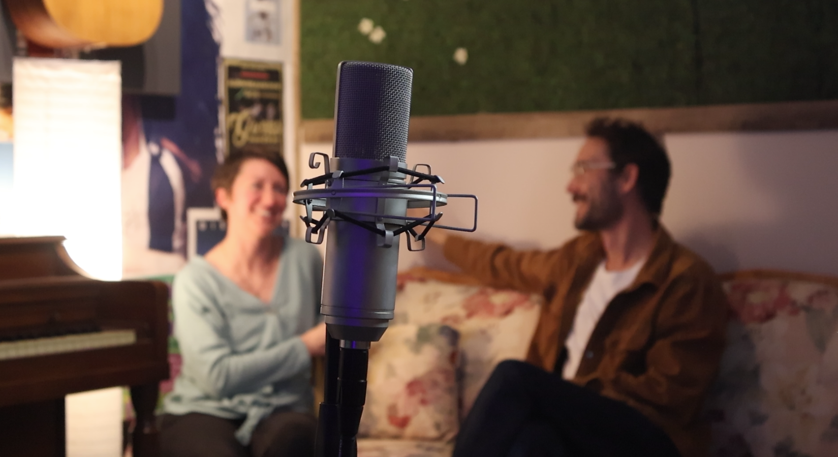 Close-up of a recording microphone in a cozy, music-themed room with two people smiling and chatting in the background.