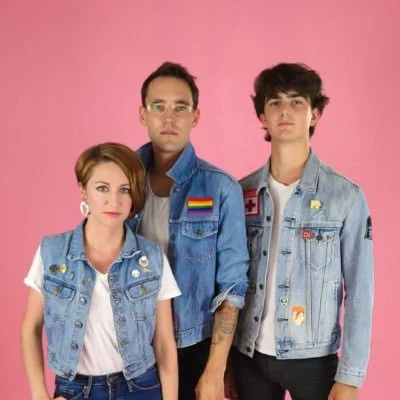 Three young adults standing against a pink background, wearing denim jackets with various patches and pins.