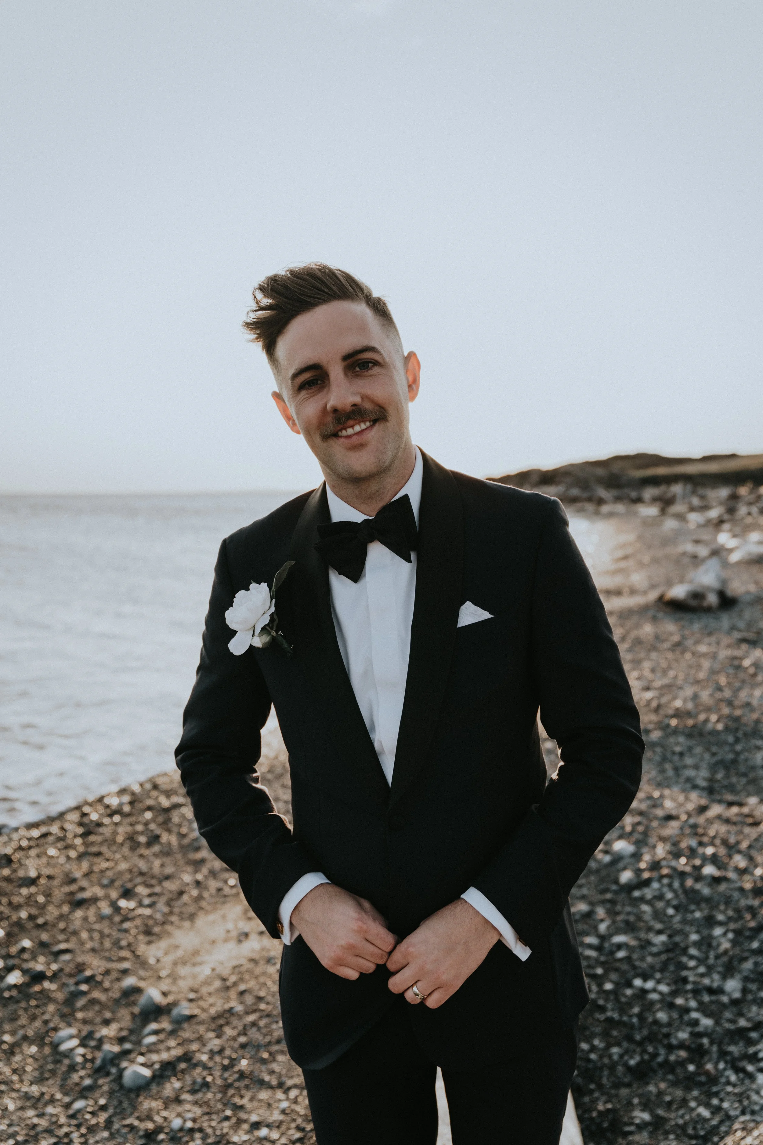 A groom in a black tuxedo with a bow tie, standing on a pebble beach near the water at sunset, smiling at the camera.
