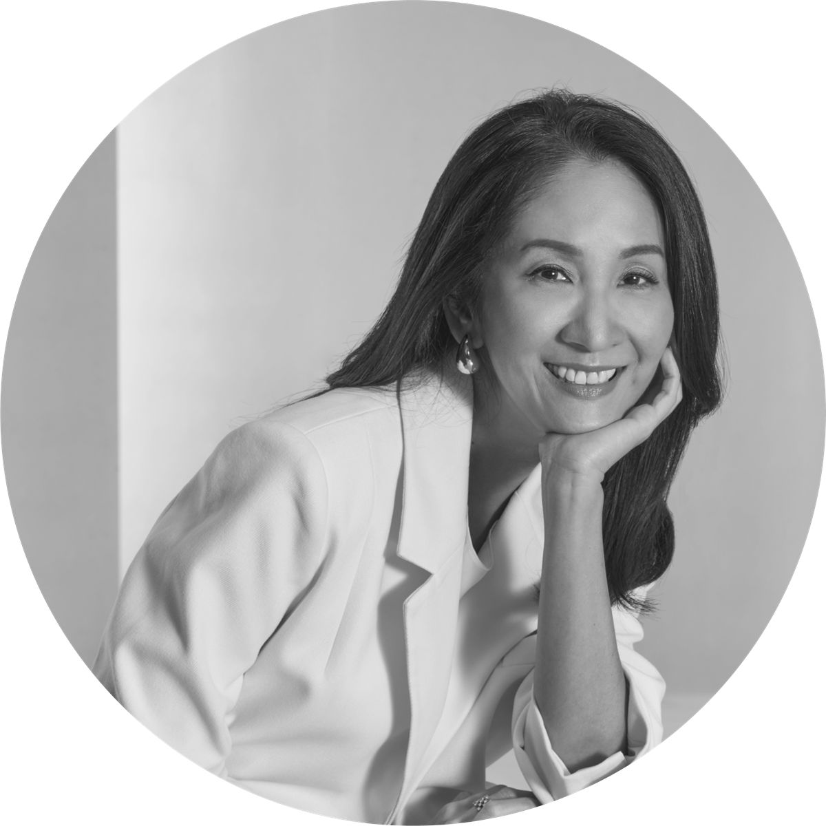 Black and white portrait of a smiling woman with straight hair, wearing earrings and a white blazer, resting her head on her hand.