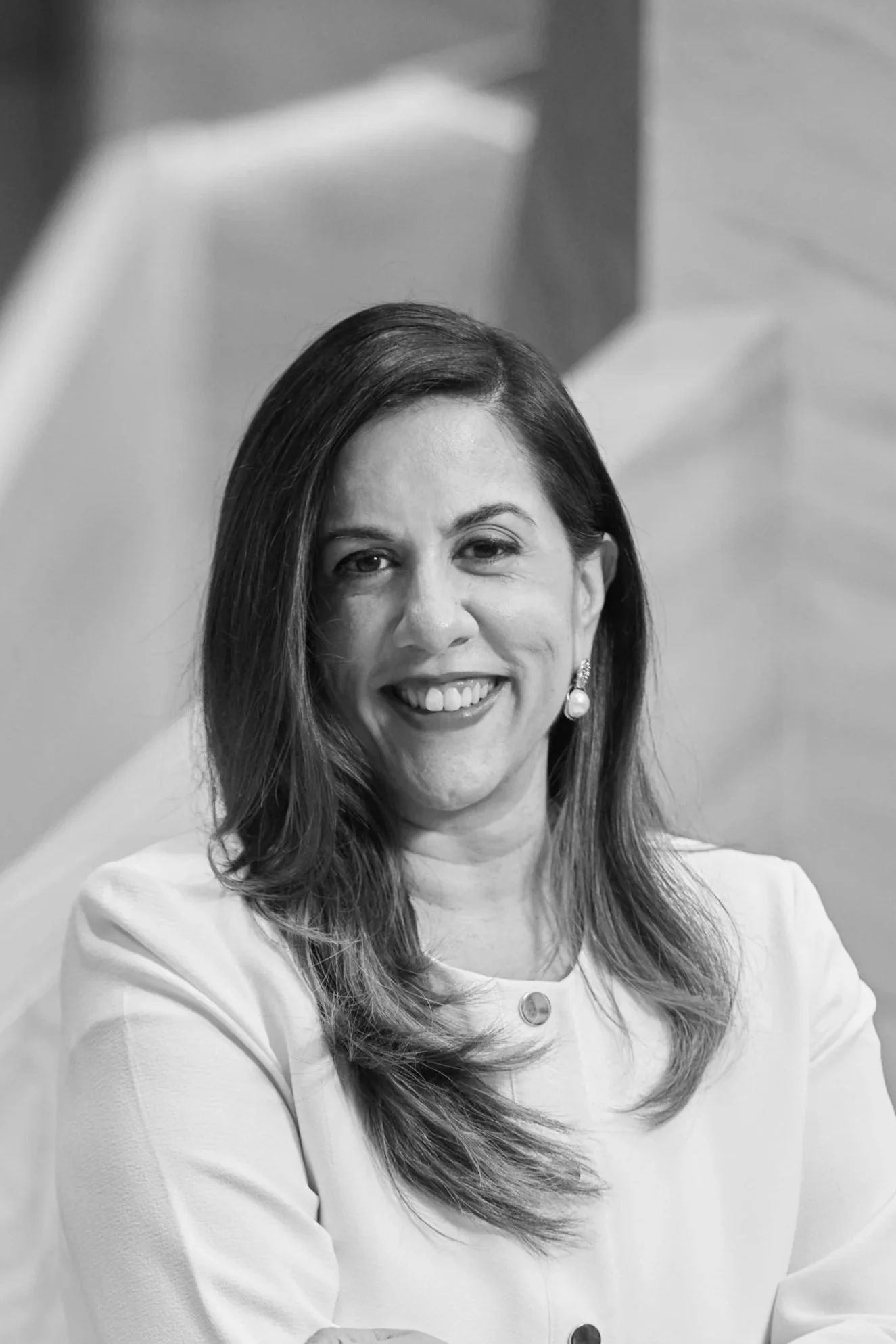 A smiling woman with long hair wearing a white top and pearl earrings.