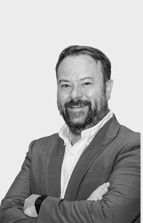 Black and white portrait of a man with a beard, smiling, wearing a suit jacket and a white shirt, with arms crossed, standing against a plain background.