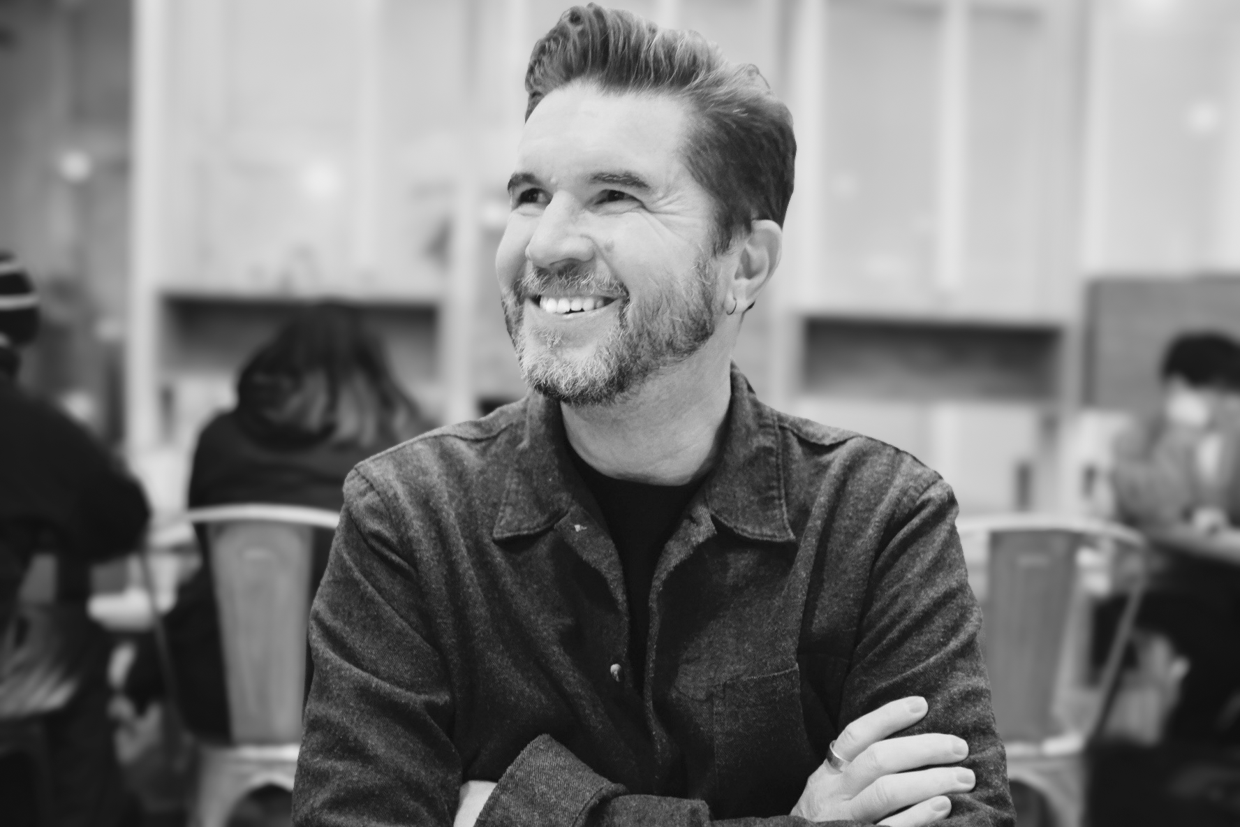 A smiling man with a beard and slicked-back hair sitting at a table with his arms crossed, in a room with other people in the background.
