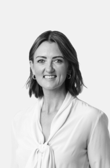 A black and white portrait of a woman with shoulder-length hair, smiling slightly, wearing a white blouse with a bow tie at the neck and hoop earrings.