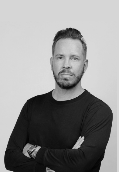 Black and white portrait of a man with short hair and a beard, wearing a long sleeve black shirt and a watch, standing against a plain light background.