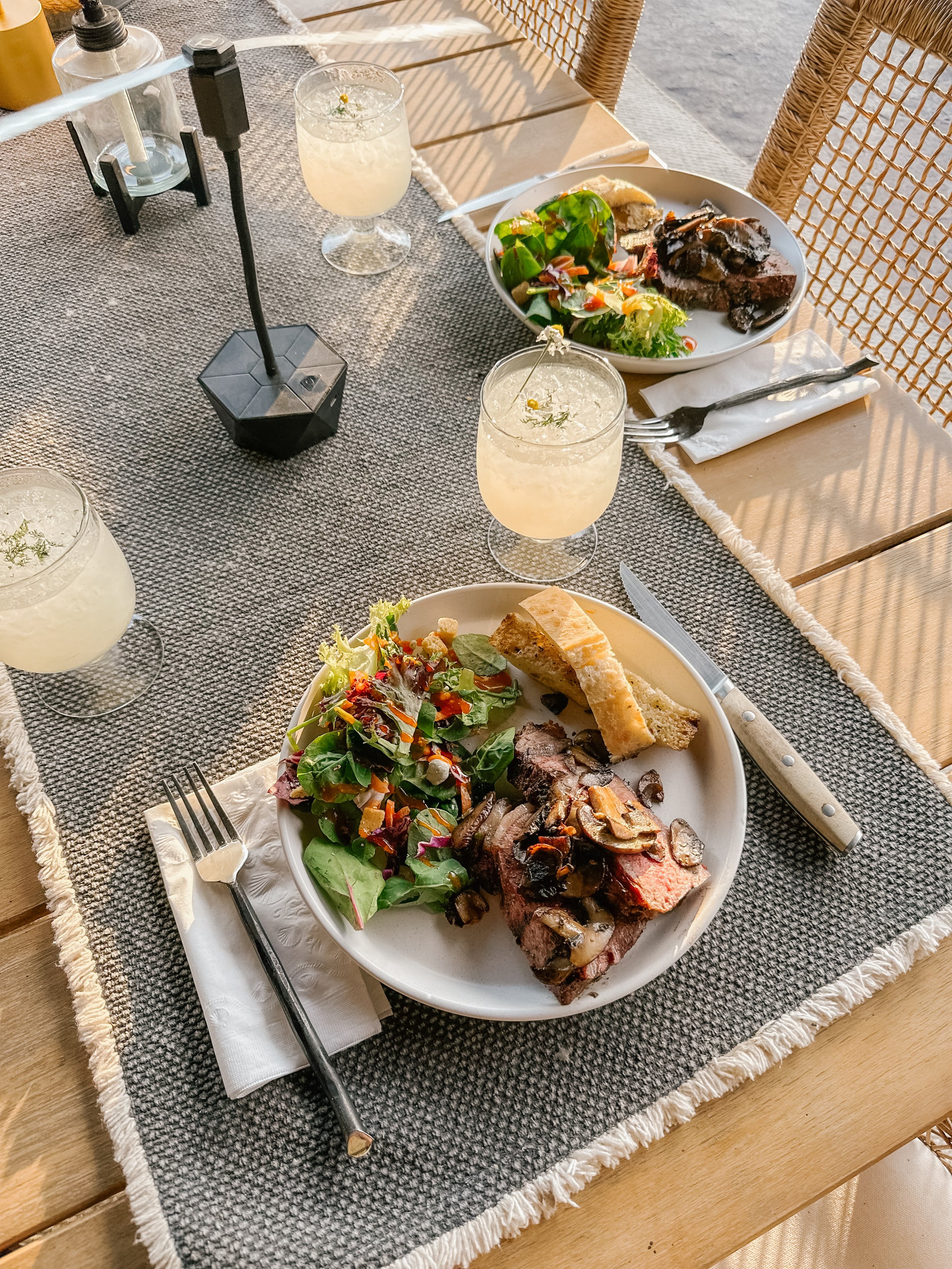 A table set for three with plates of beef, salad, and bread, two glasses of lemonade, and a candle in a modern, outdoor setting.