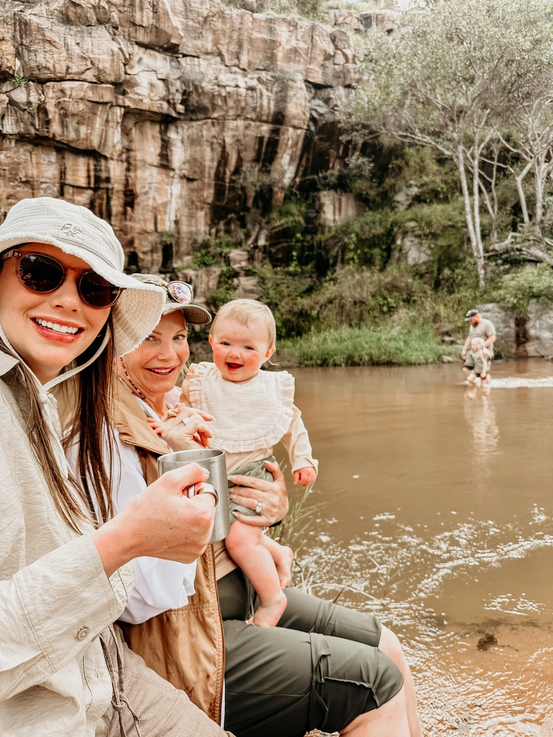 Smiling women and a baby sitting by a river with a rocky cliff in the background, one woman holding a metal cup, others in outdoor clothing, enjoying a day by the water.