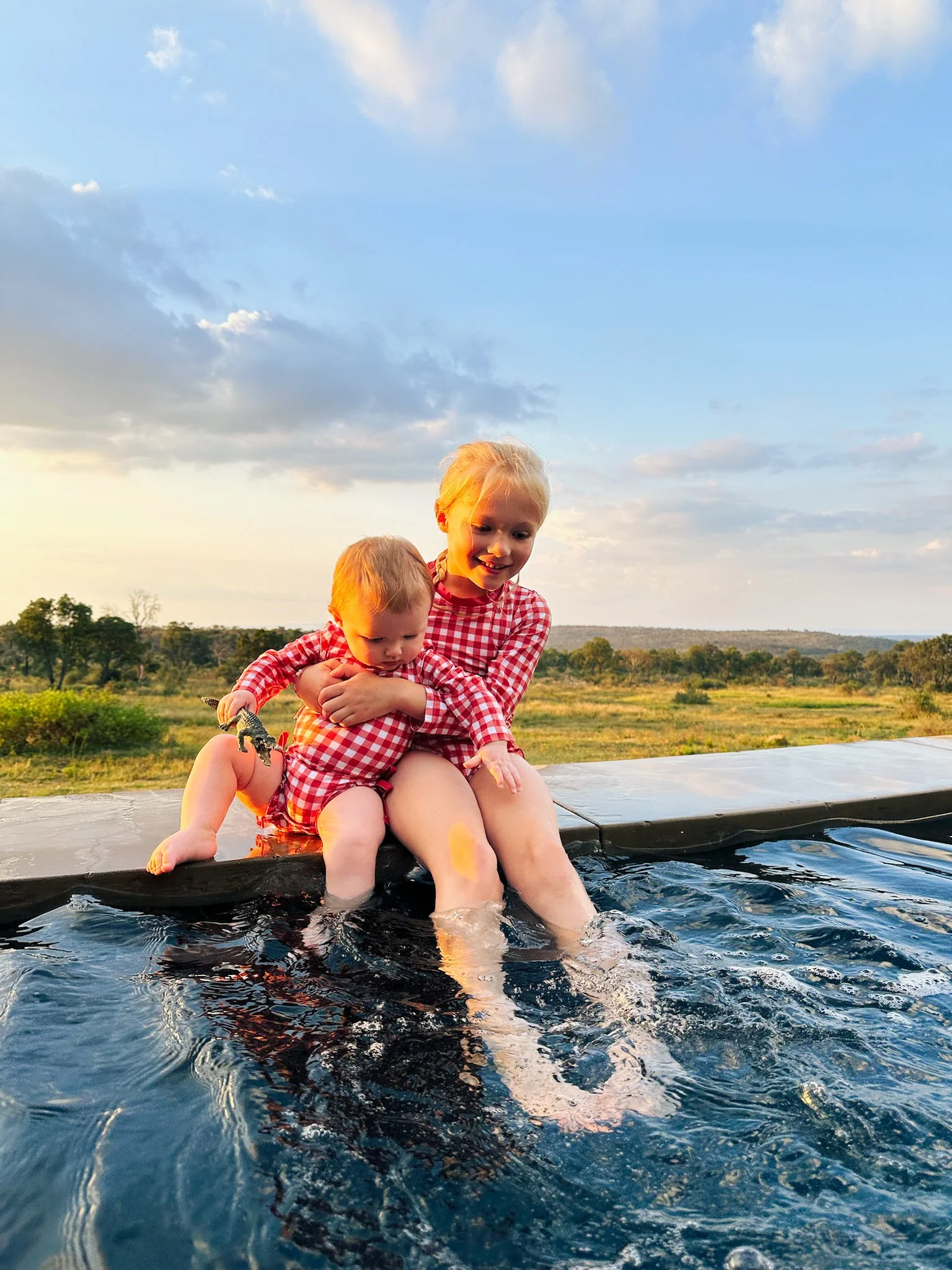 Two young children wearing matching red and white checkered outfits sit on the edge of a pool, with one child splashing in the water and the other holding a small toy, set against an open field and sky at sunset.