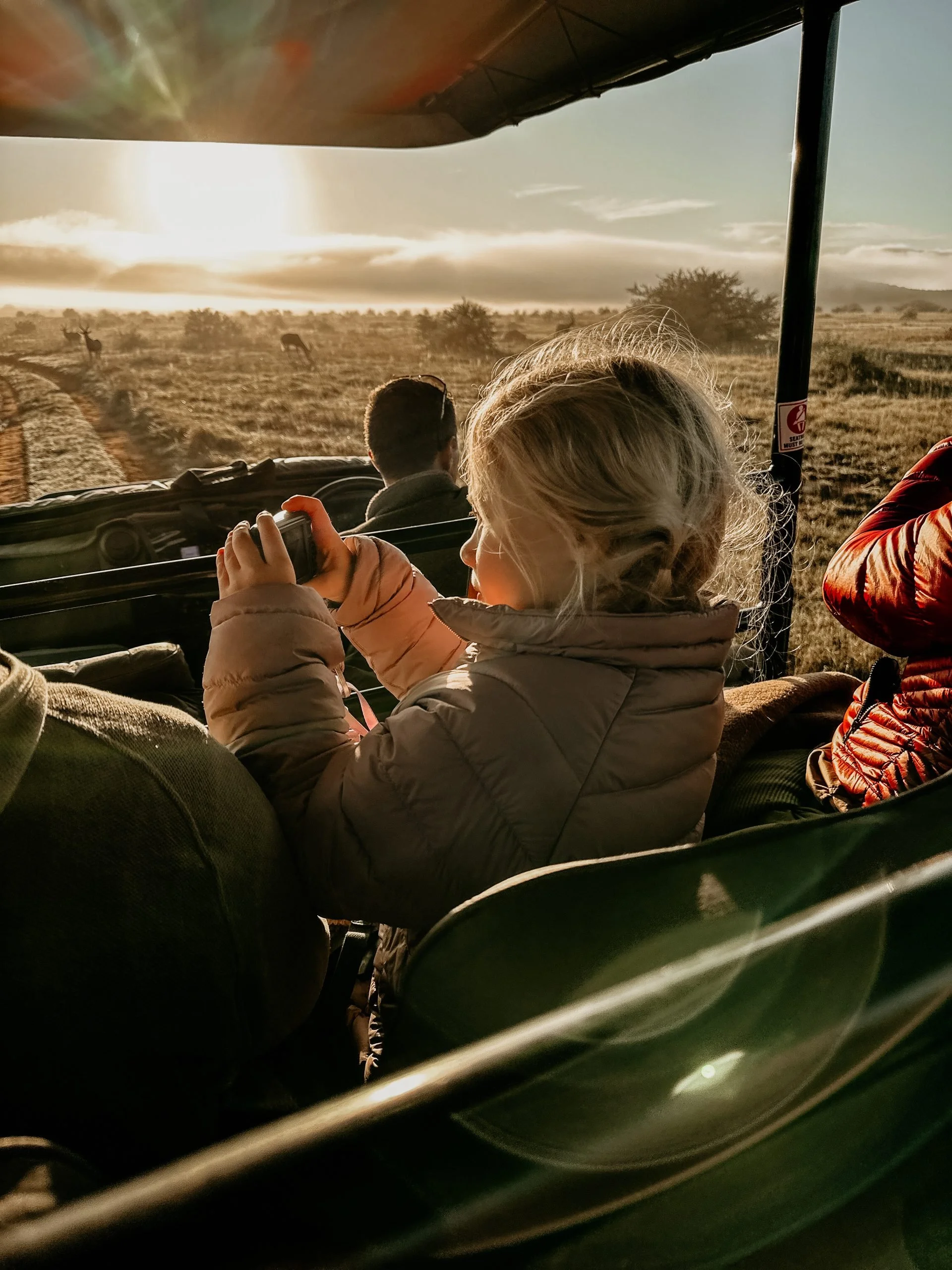 A young girl with blonde hair wearing a tan jacket seated in a safari vehicle, looking out at a landscape with a dirt path, open grassland, and a few animals in the distance, during sunset or sunrise.