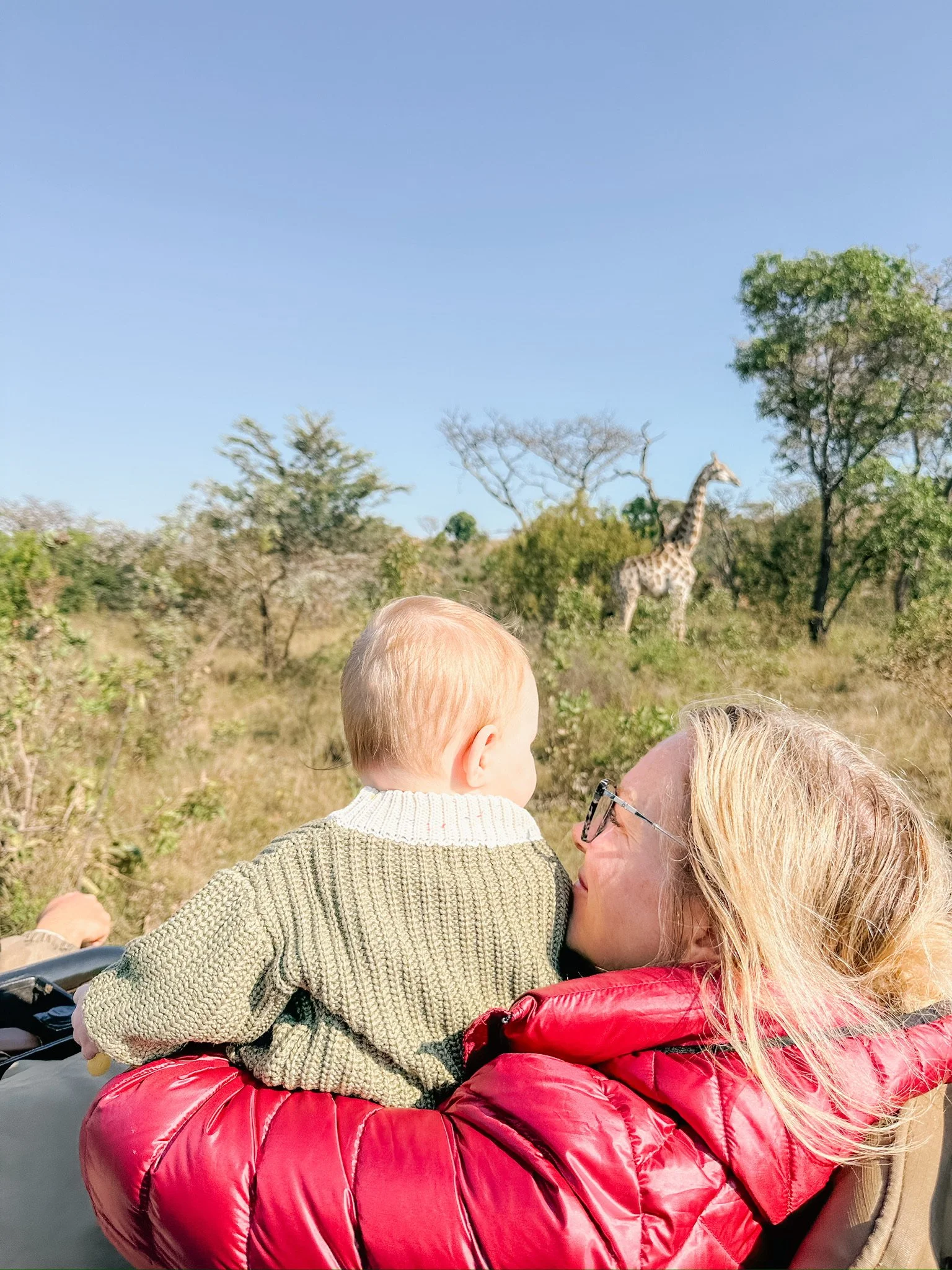 A woman holding a young child in a safari park with a giraffe in the background.