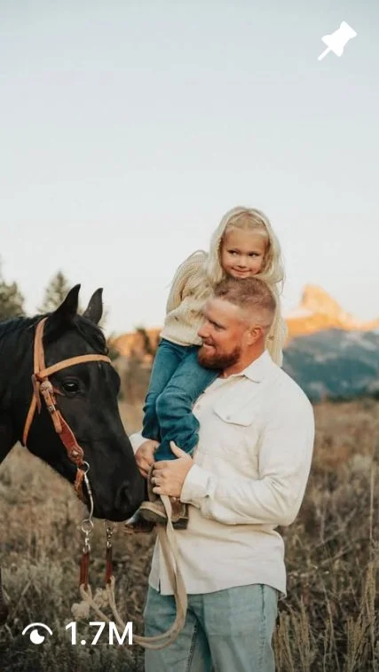 A man holding a young girl on his shoulders next to a black horse in a rural outdoor setting at sunset.