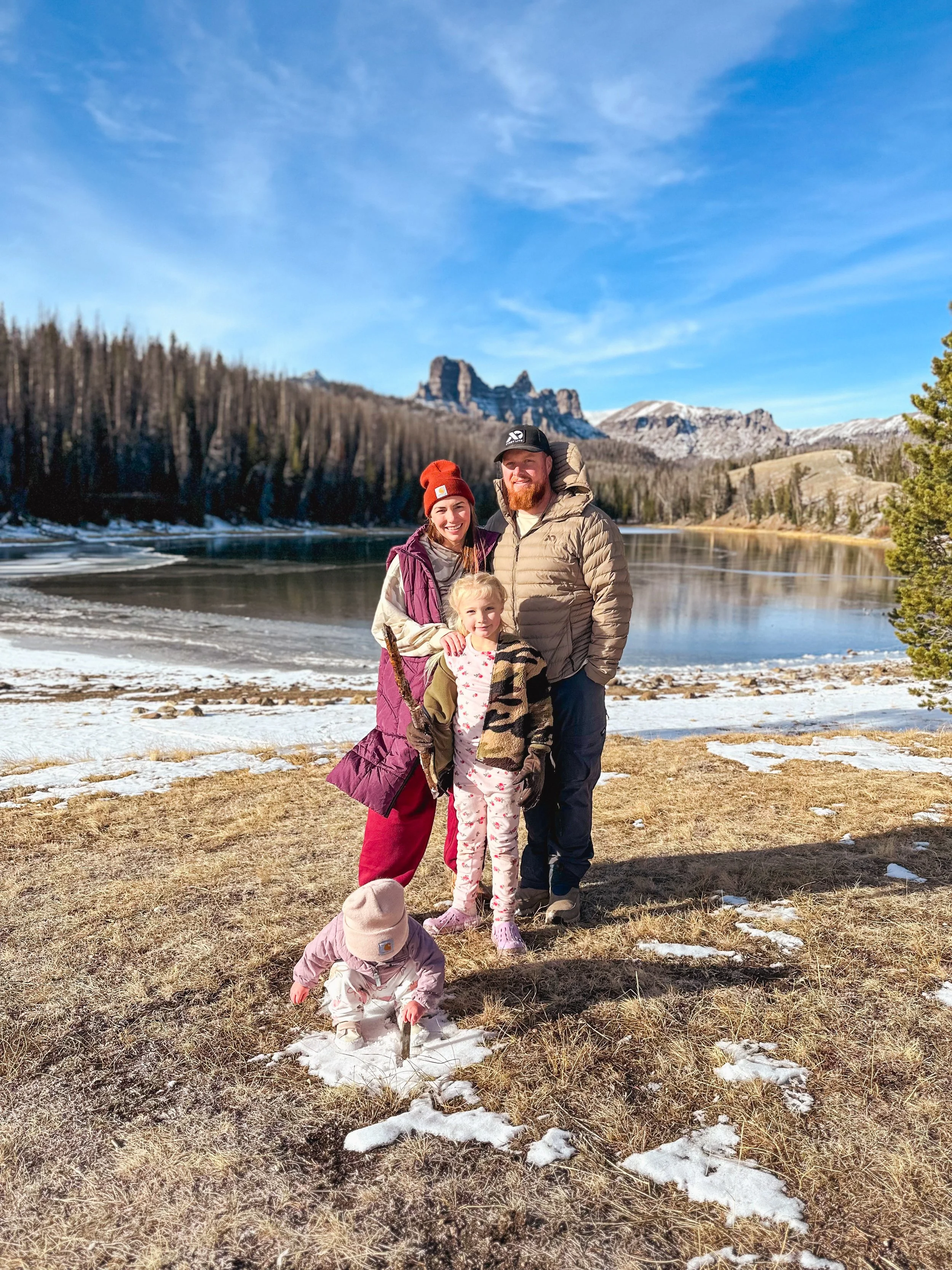 Family of four standing outdoors by a partially frozen lake with mountains in the background, dressed in winter clothing.
