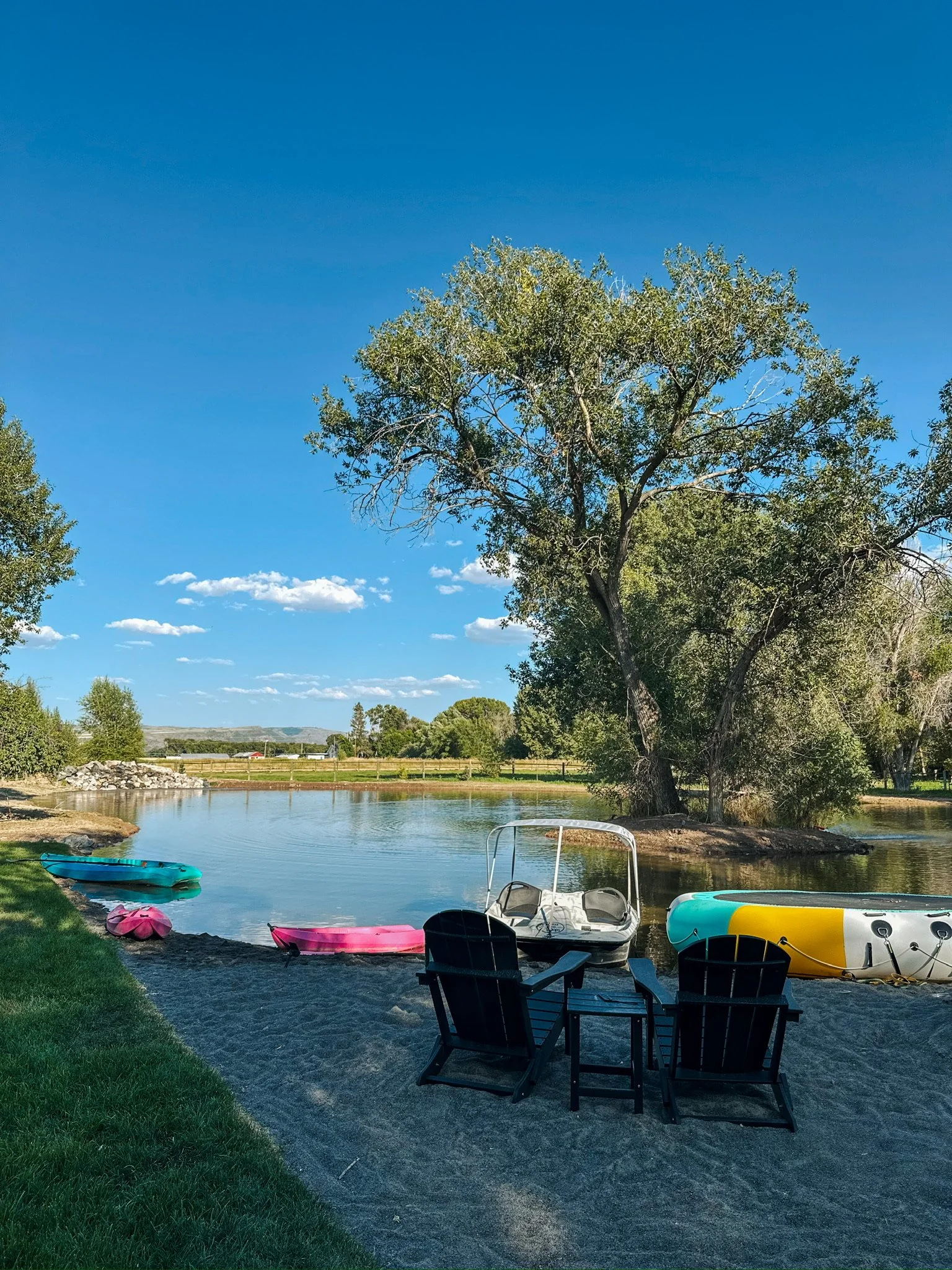 Two black Adirondack chairs and a small table on a sandy beach next to a river with paddle boats and a large tree under a blue sky with clouds.