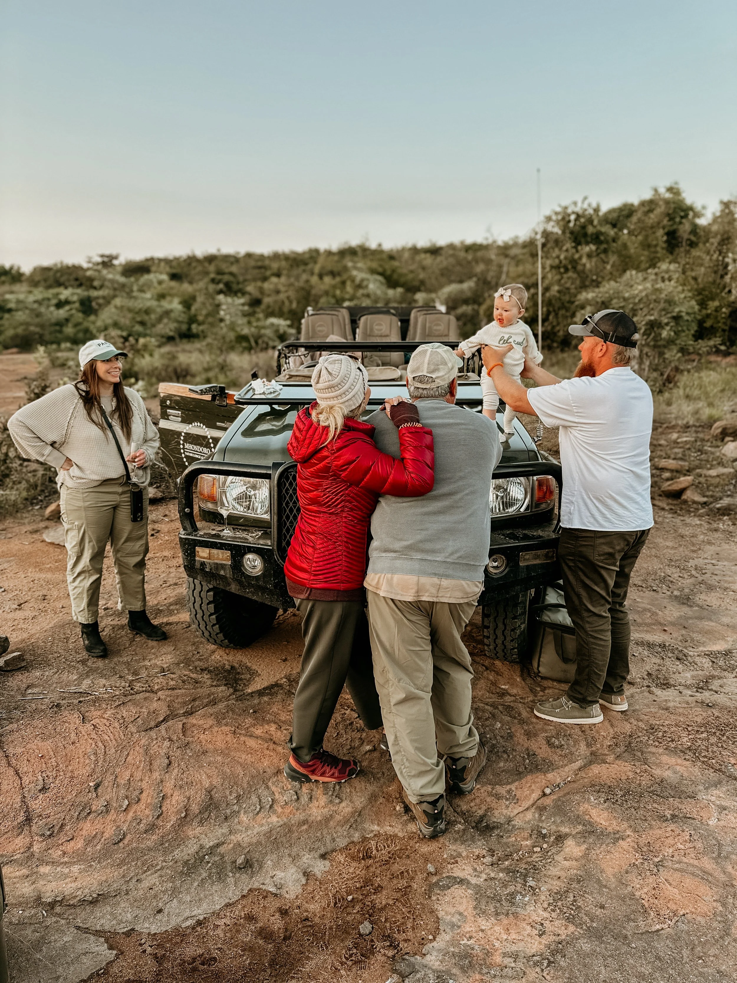 Family enjoying time together outdoors near a black off-road vehicle, with a young girl and adults, in a natural landscape.
