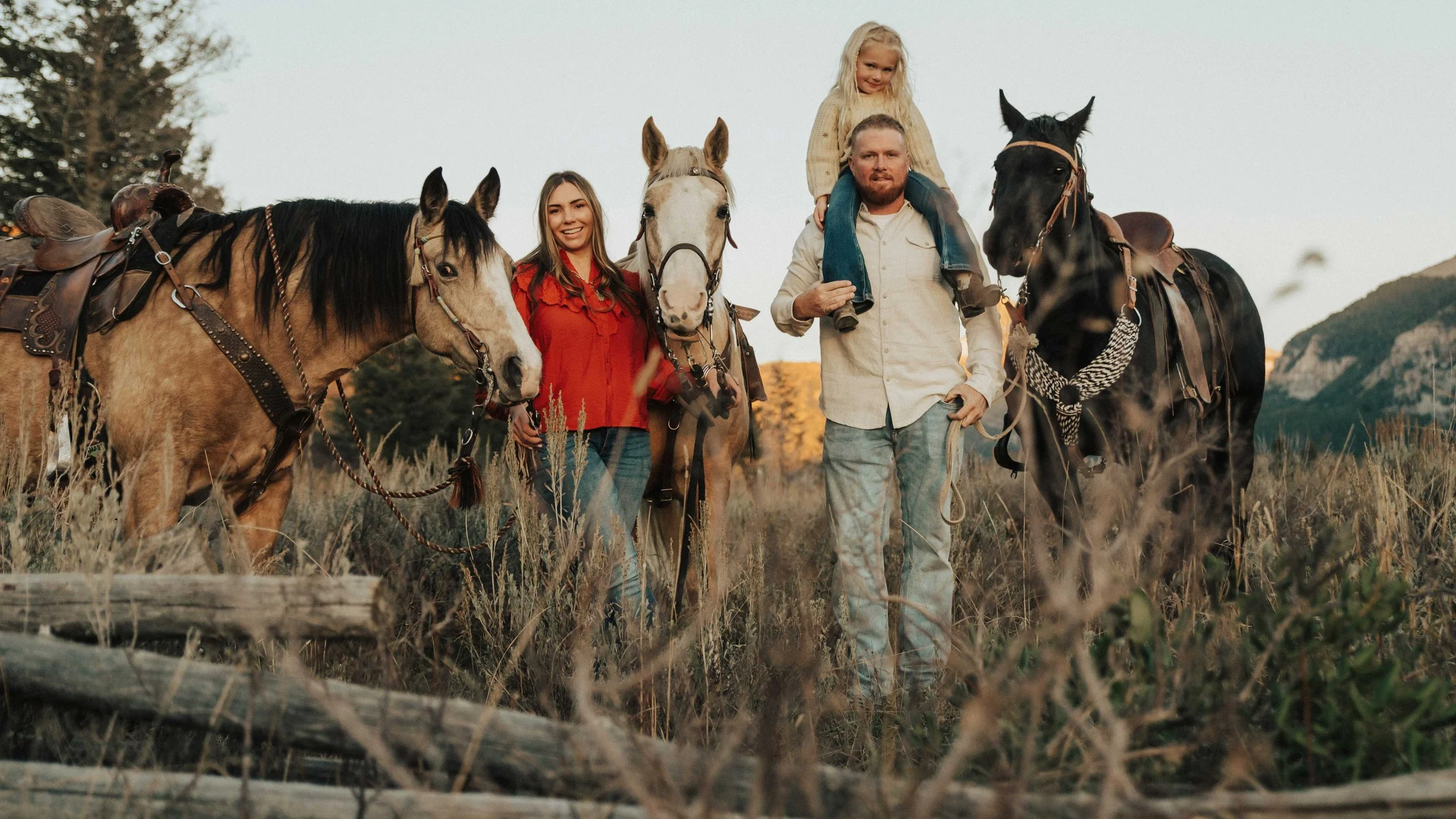 A family of five with three children standing in a field with three horses during sunset, mountains in the background.