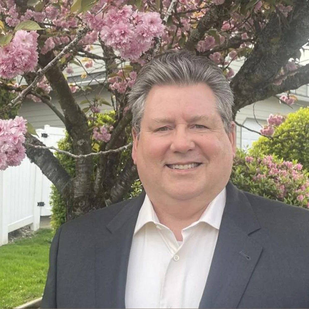 Mark Palmer standing in a suit standing outside in front of a blossoming pink tree with white fence and house in the background.