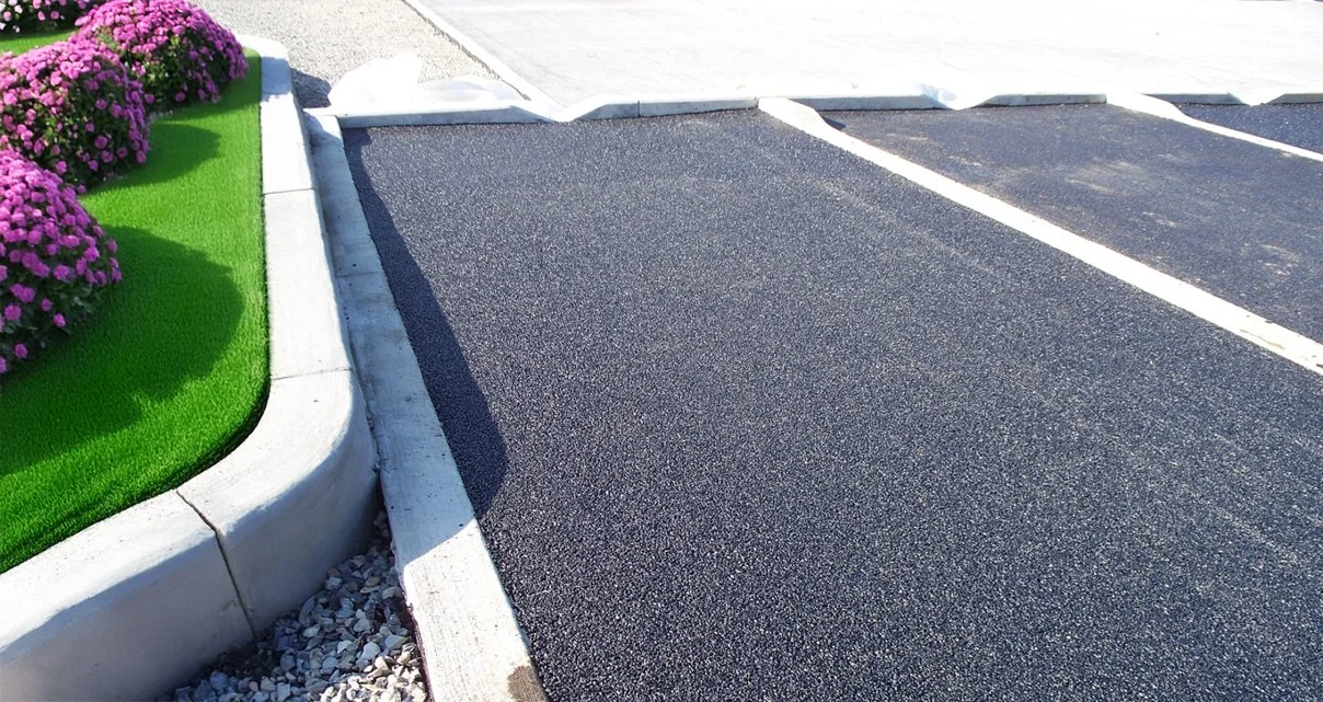 A parking lot with porous pavement and white painted parking lines, adjacent to a green grass patch with purple flowers and a white concrete curb.