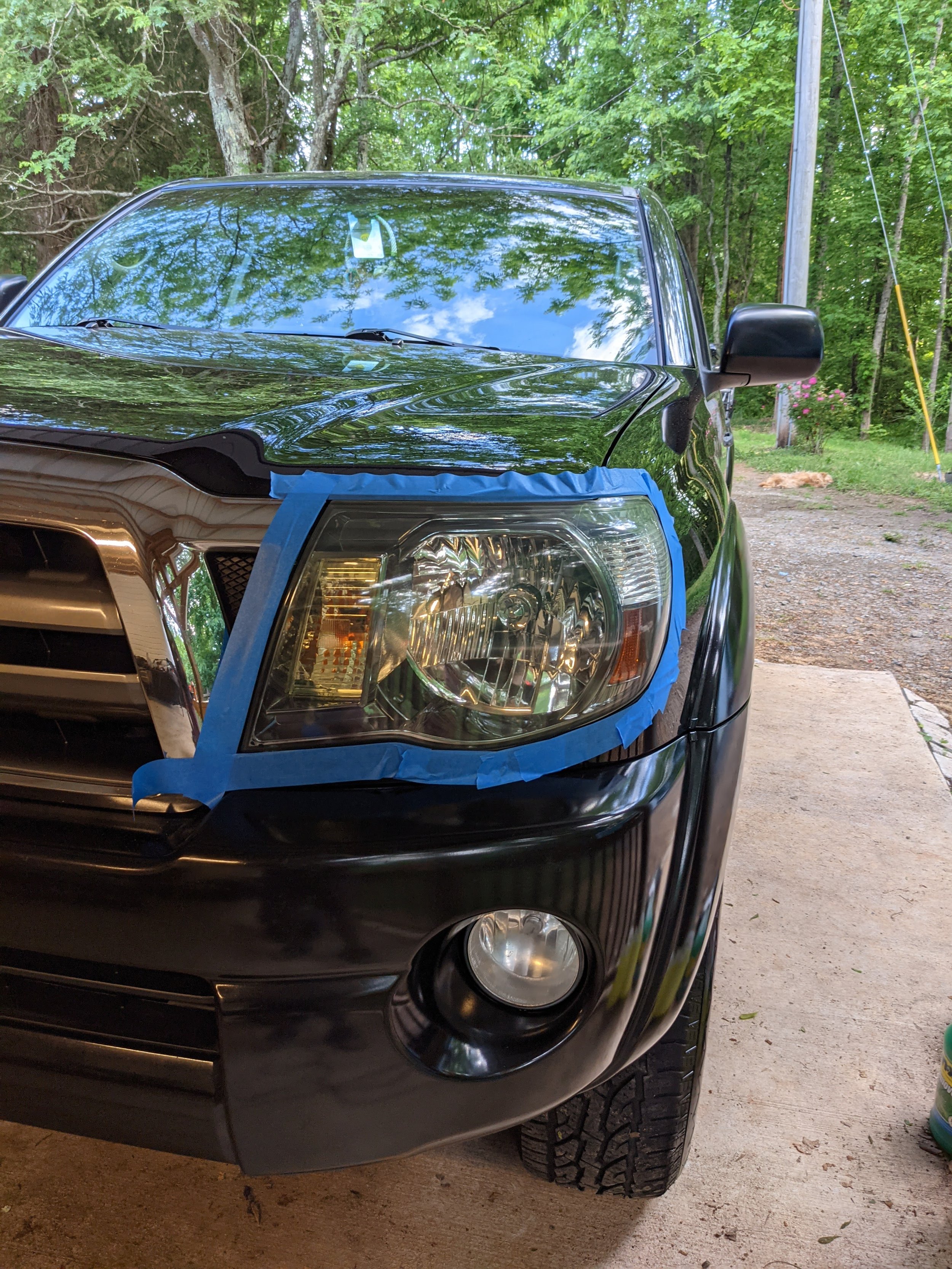 Front of a black vehicle with a damaged headlight, masked with blue painter's tape, parked outdoors on a concrete driveway, surrounded by trees and greenery.