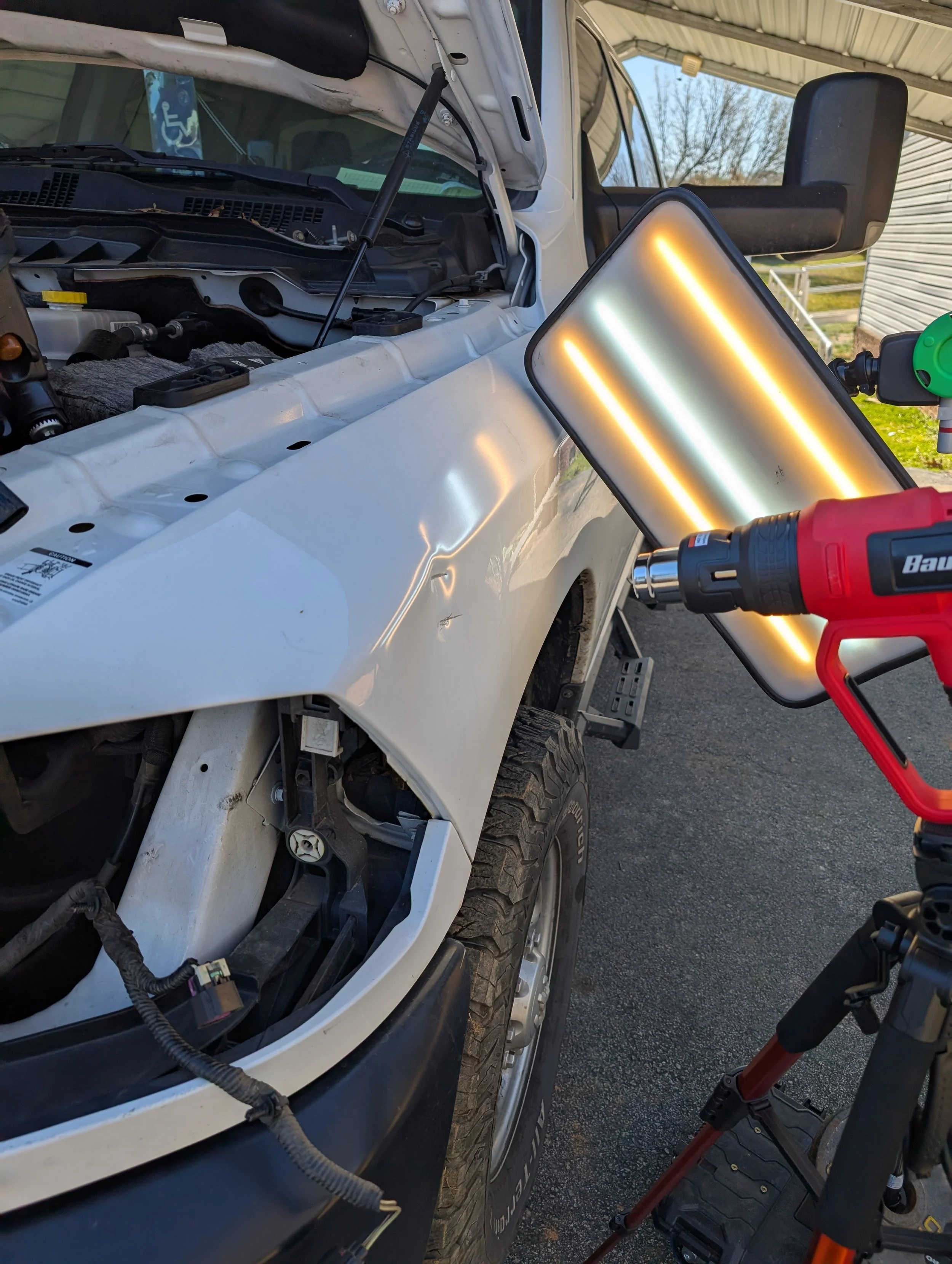 A white vehicle with its hood open, undergoing body work or repair, with a red heat gun directed at a metal panel and a work light illuminating the area.