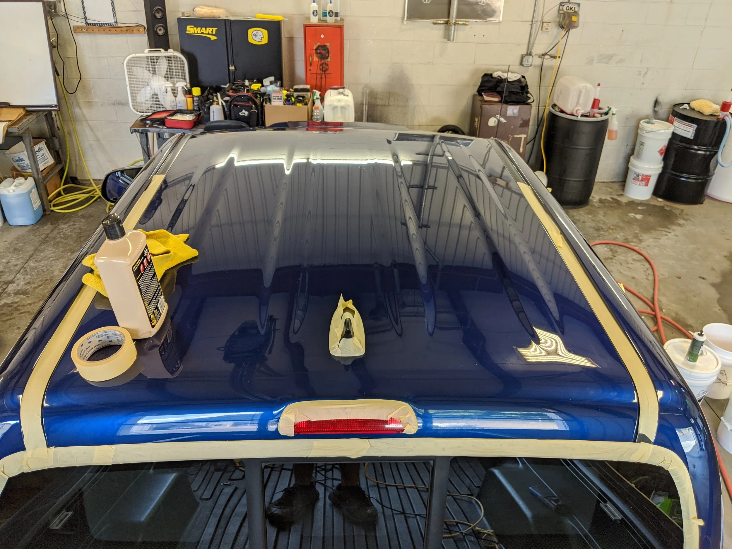 The image shows a blue car in a garage with masking tape and supplies on its rear windshield, prepared for painting or detailing.