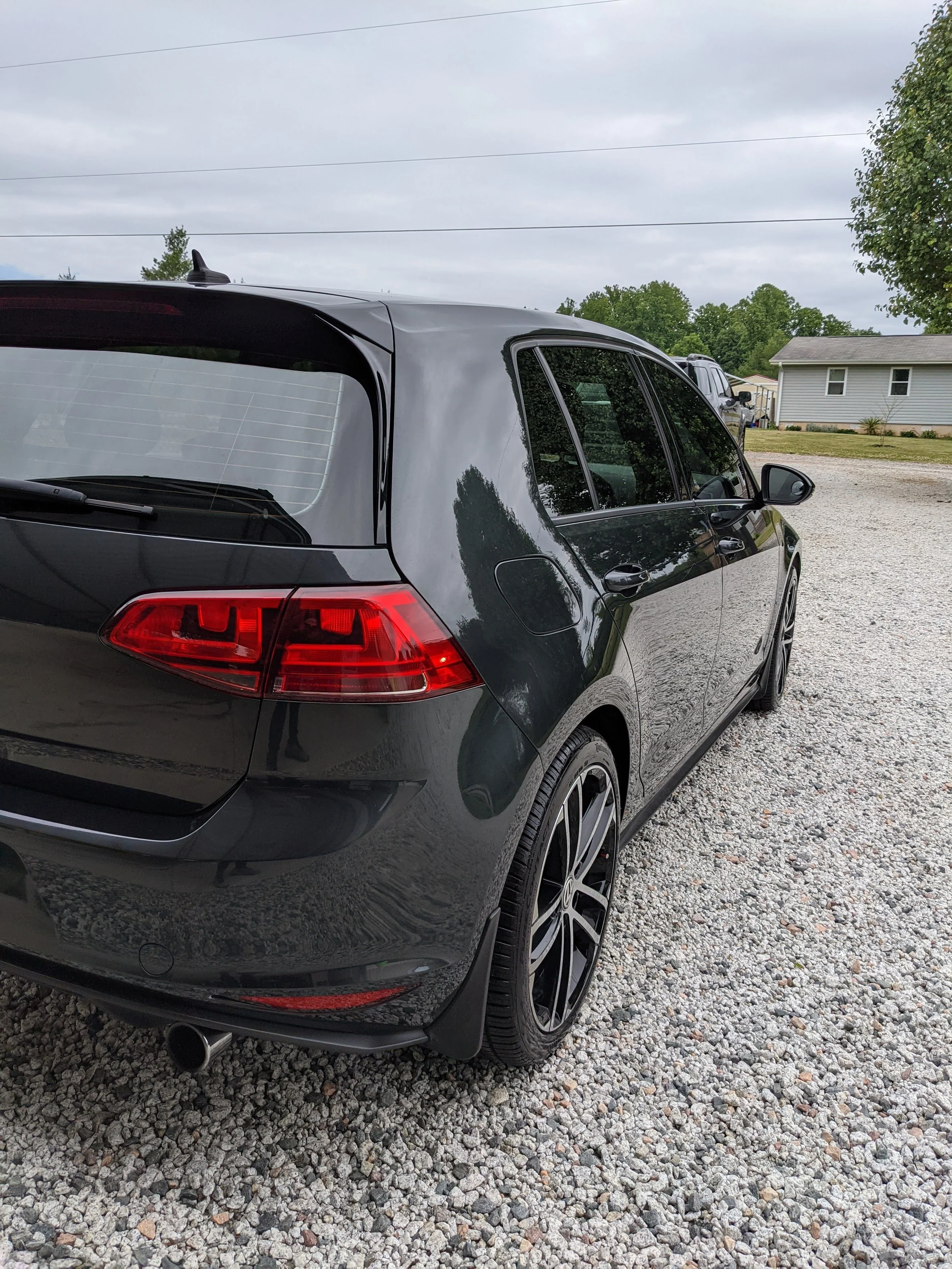 Black hatchback car parked on a gravel lot, with trees and a house in background.